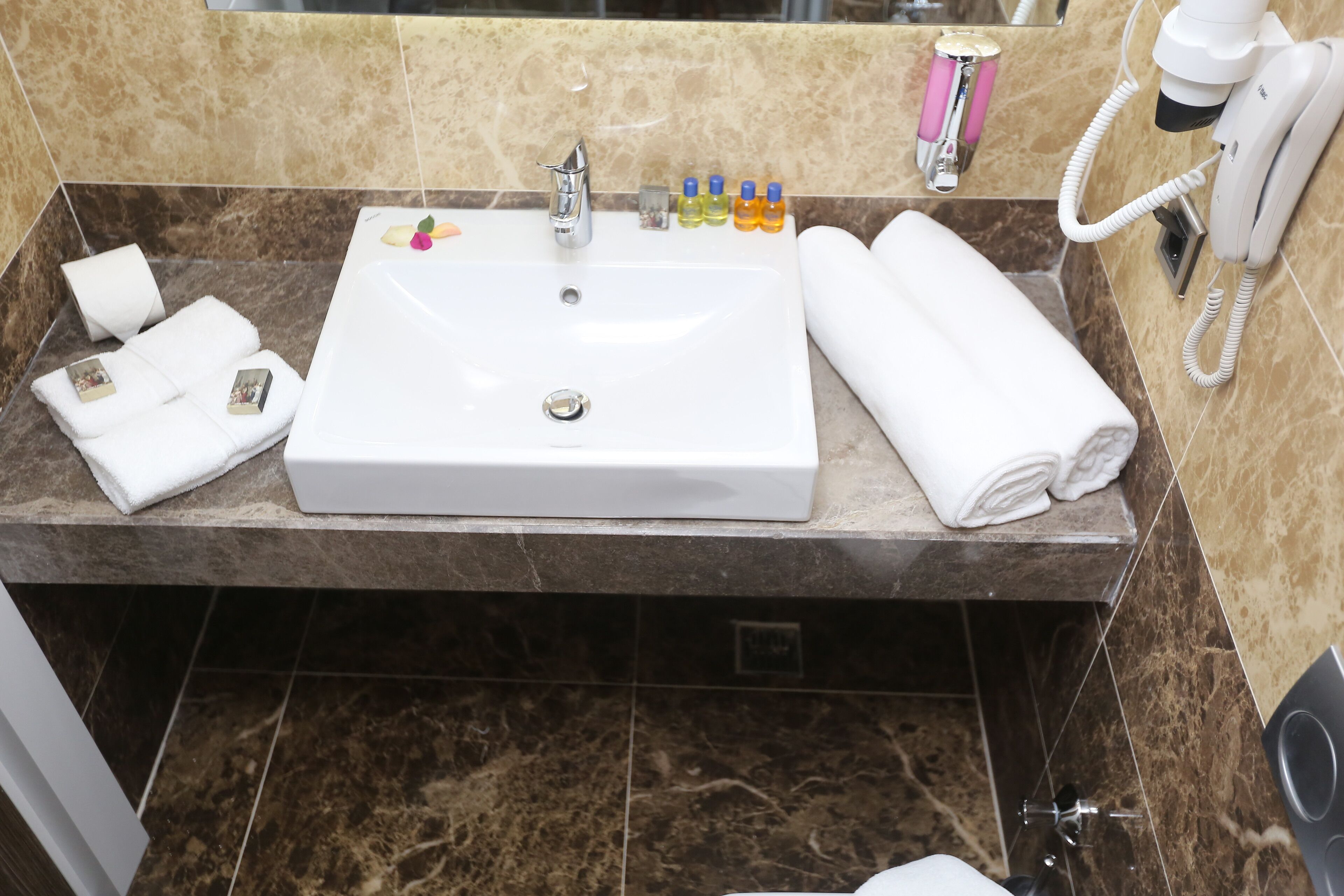 Un baño con un lavamanos blanco, baldosas de mármol, un espejo, un secador de pelo y artículos de aseo.
