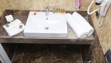 Un baño con un lavamanos blanco, baldosas de mármol, un espejo, un secador de pelo y artículos de aseo.