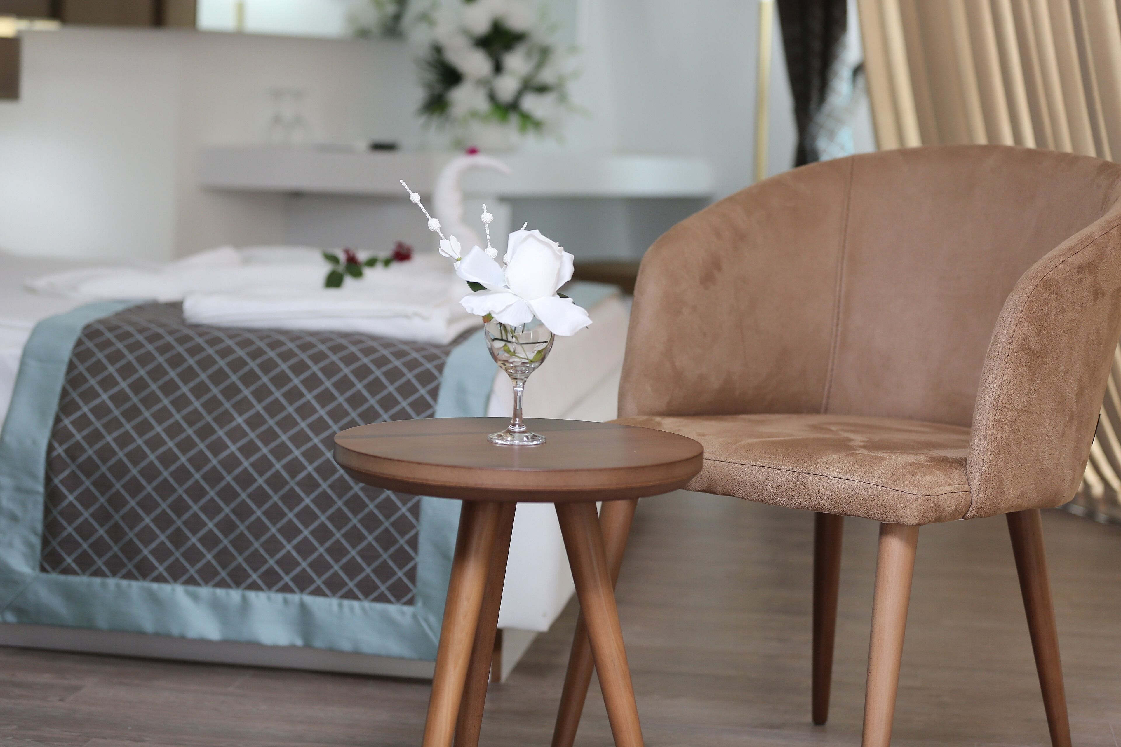 Un dormitorio con una cama, una mesa redonda de madera con un florero de cristal y flores, y una silla tapizada marrón.