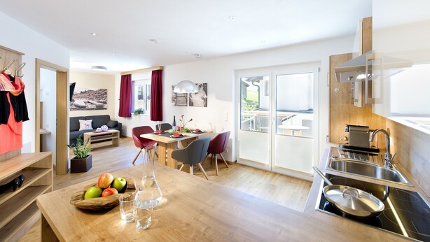 A modern kitchen with a built-in stove, a dining area with a wooden table and chairs, and a living area with a sofa and a coffee table. - Dorfjuwel Appartements (Wildschönau)