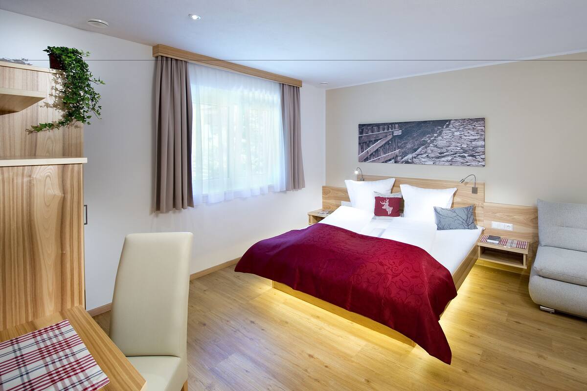 A modern hotel room with a wooden bed, a red throw blanket, a grey armchair, and a wooden nightstand.