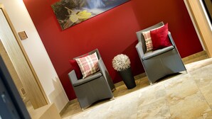 A reception area with two grey armchairs, one with a red and white checkered pillow and the other with a red pillow. A large painting of a natural scene hangs on the red wall above the chairs. - Dorfjuwel Appartements (Wildschönau)