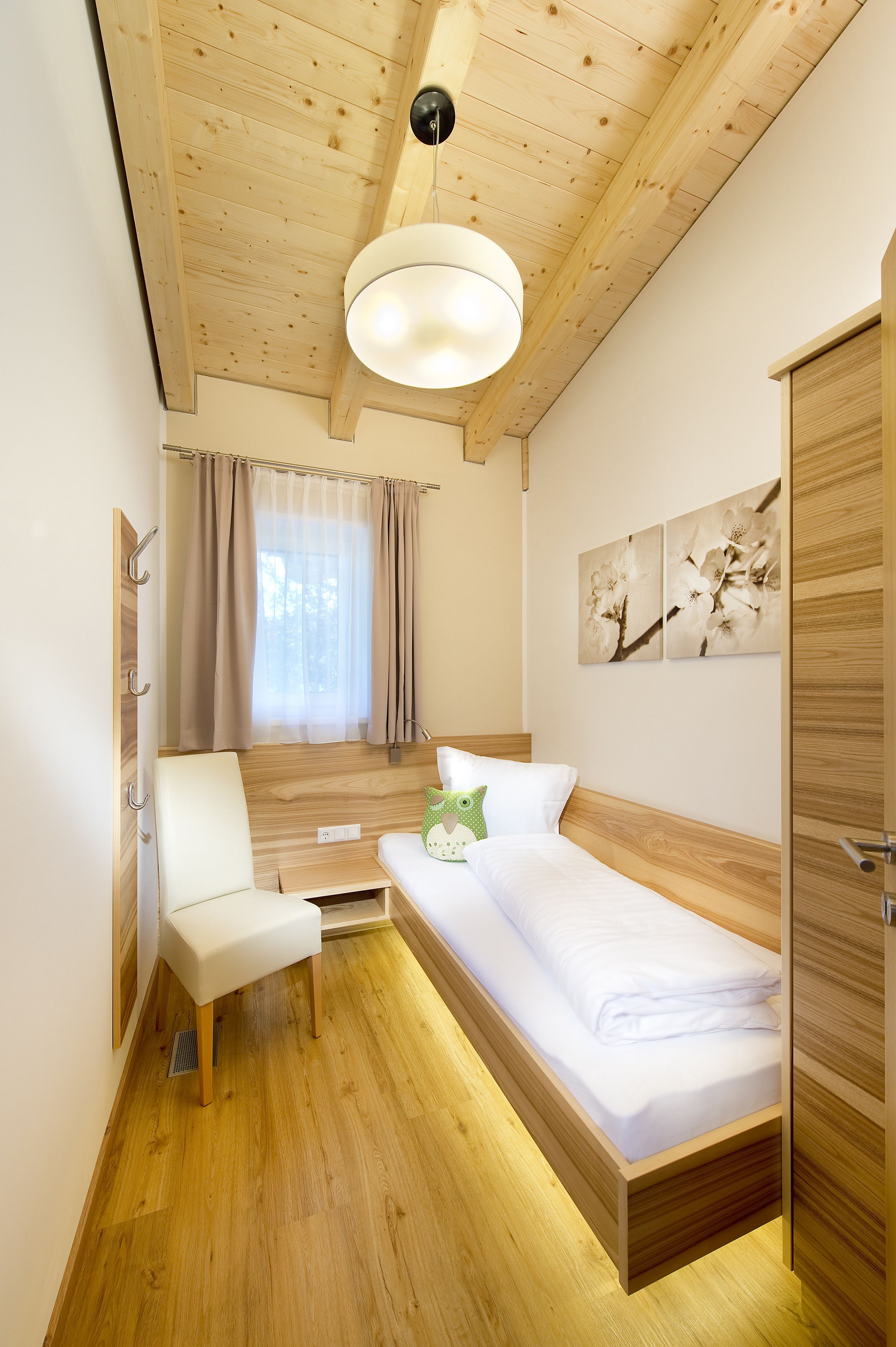 A compact bedroom with a wooden ceiling, a single bed, a chair, a small table, and a window with curtains.