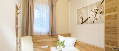 A compact bedroom with a wooden ceiling, a single bed, a chair, a small table, and a window with curtains.