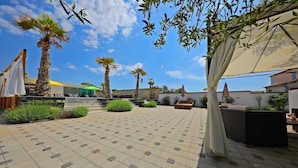 A paved outdoor area with palm trees, a covered seating area, and a building in the background. - Boutique Apartments Blitz (Bibinje)