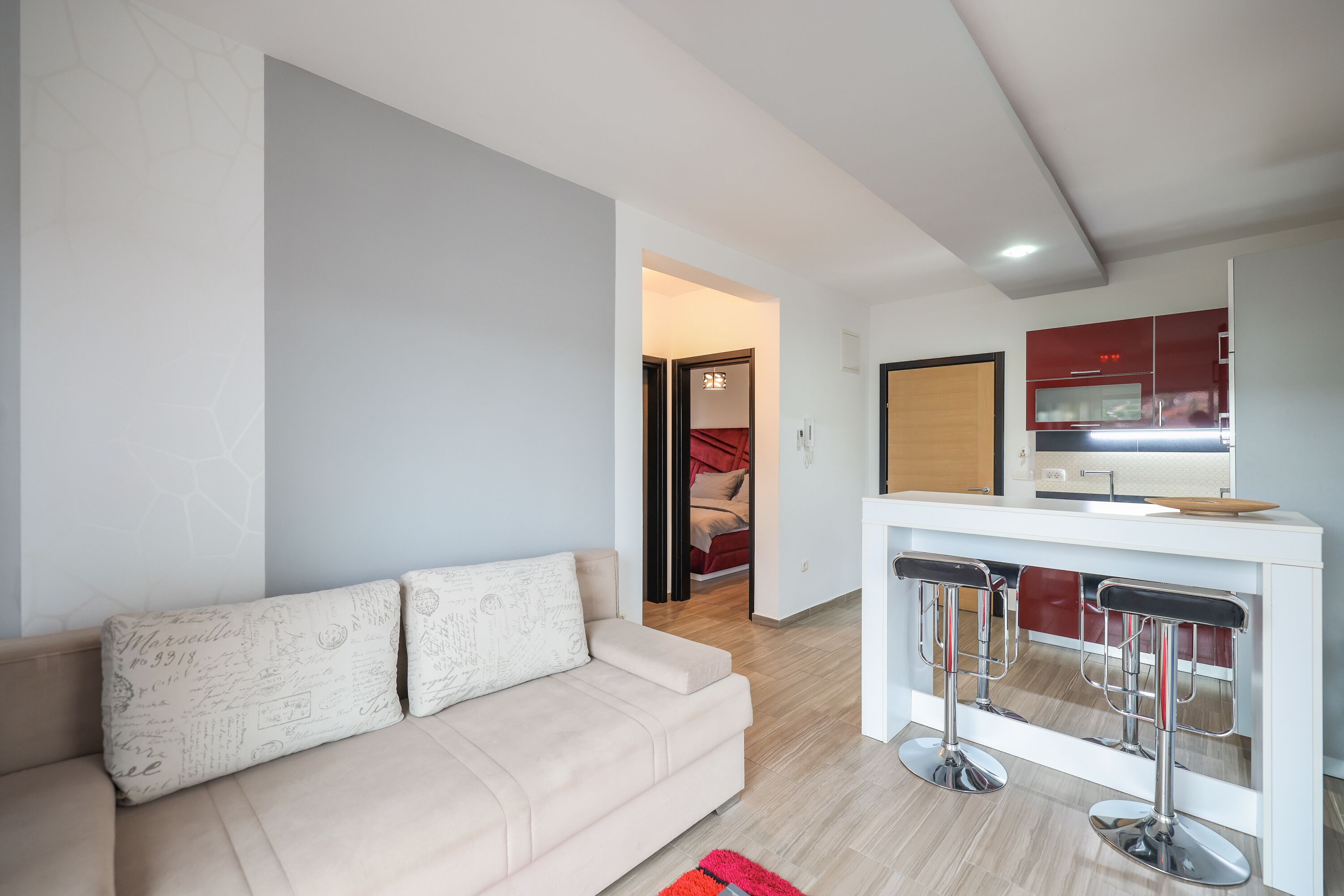 A modern living room with a beige sofa, a white coffee table, and a kitchen area with red cabinets and a white countertop.
