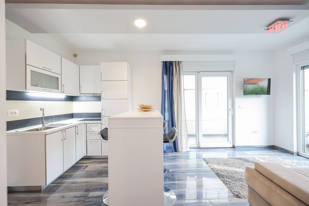Una cocina moderna con armarios blancos, un microondas y un fregadero. Hay un comedor con una mesa y sillas, y un ventanal con cortinas.