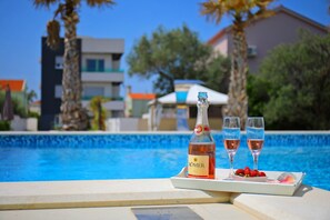 Una botella de vino RÖMER, dos copas y una bandeja con fresas y un pétalo de rosa junto a una piscina.