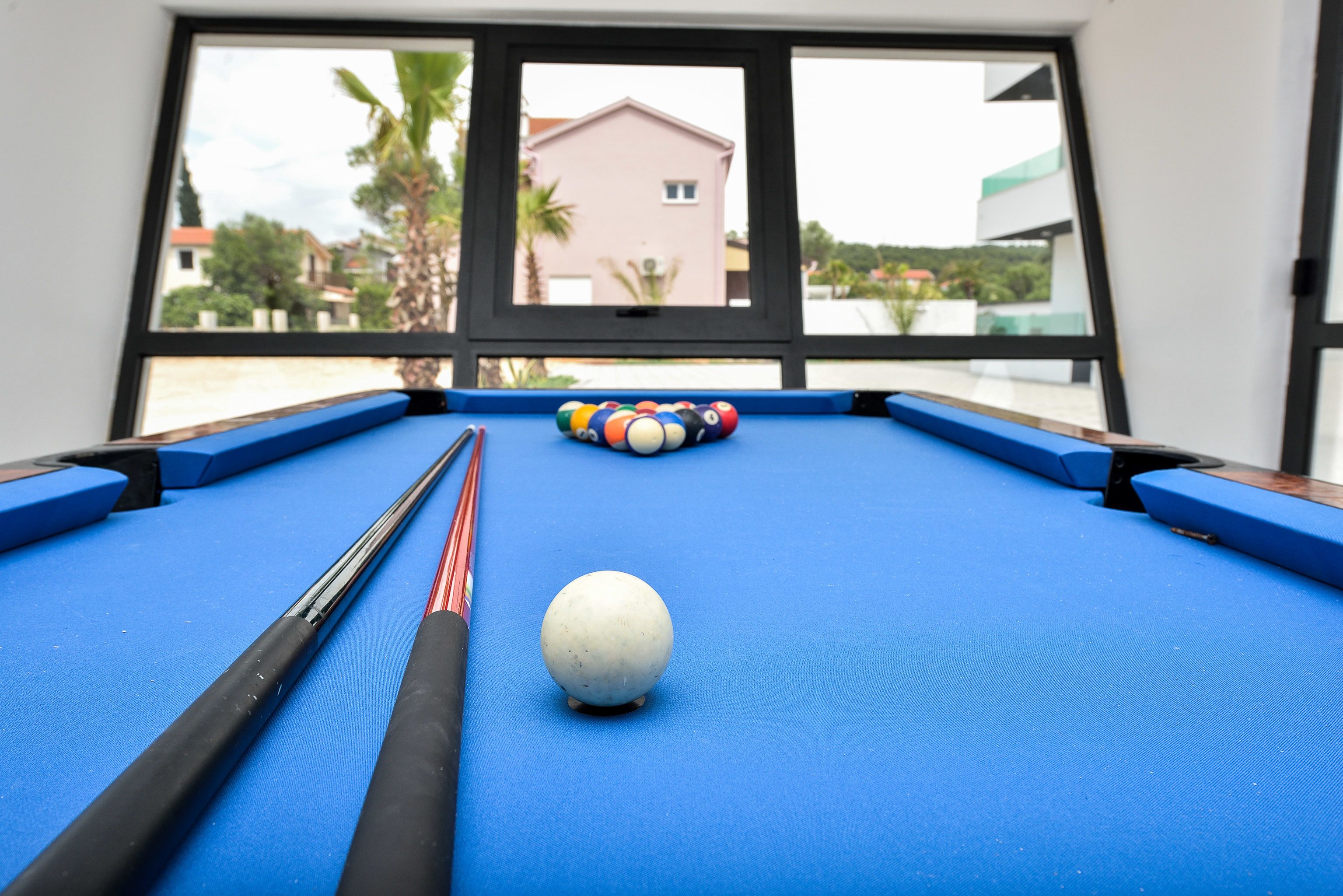 A pool table with a white cue ball and a rack of billiard balls, viewed through a window overlooking a building and palm trees.