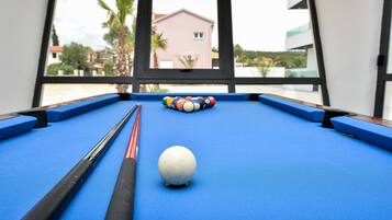 A pool table with a white cue ball and a rack of billiard balls, viewed through a window overlooking a building and palm trees.