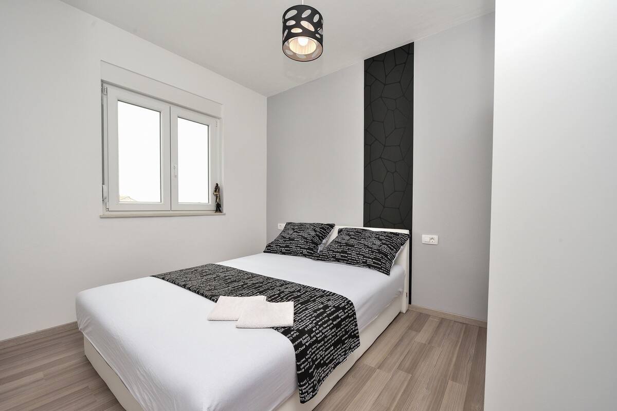 Un dormitorio con cama, ventana, lámpara fijada a la pared y pared con textura.