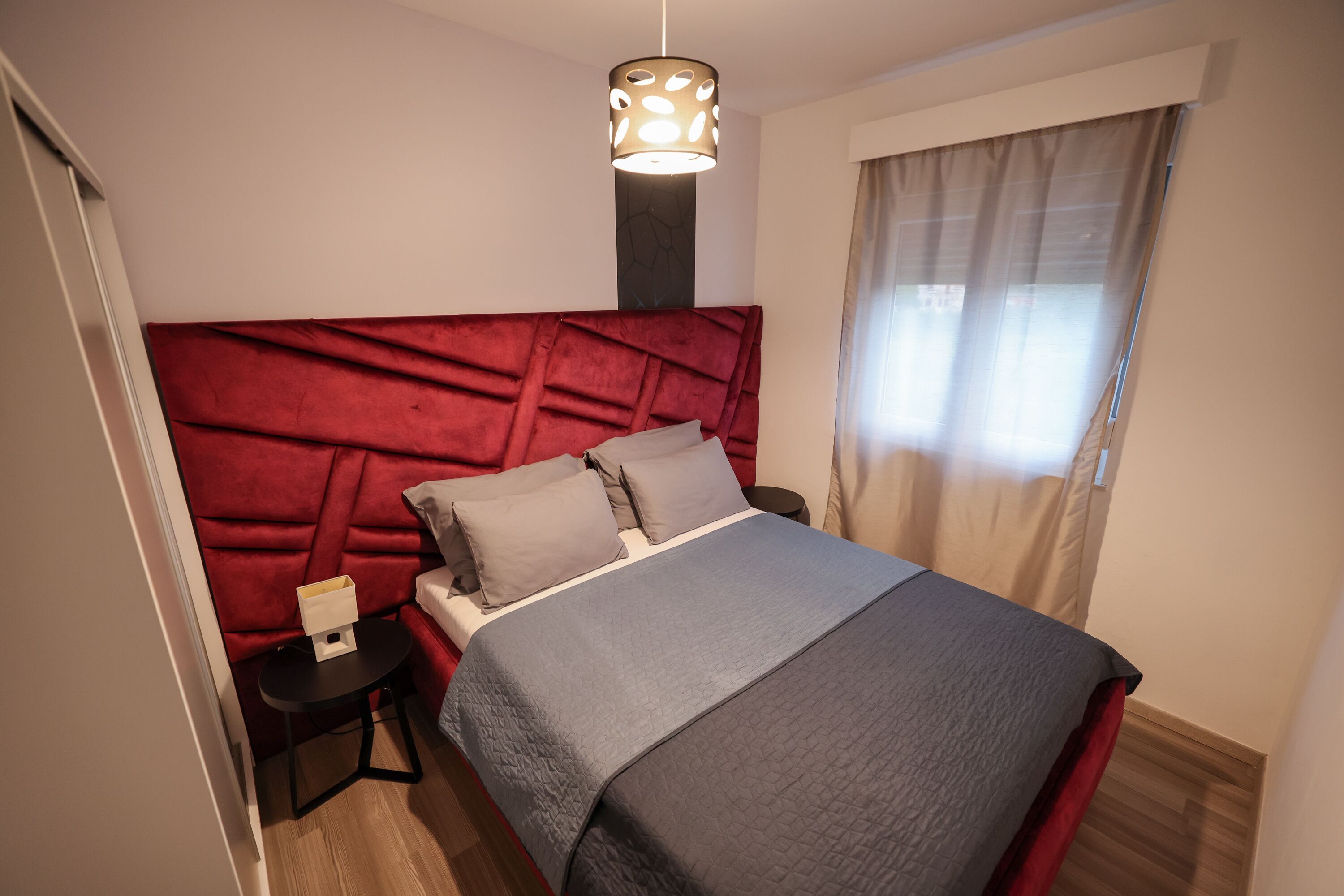 A bedroom with a red headboard, a grey bedspread, a bedside table with a lamp, and a window with curtains.