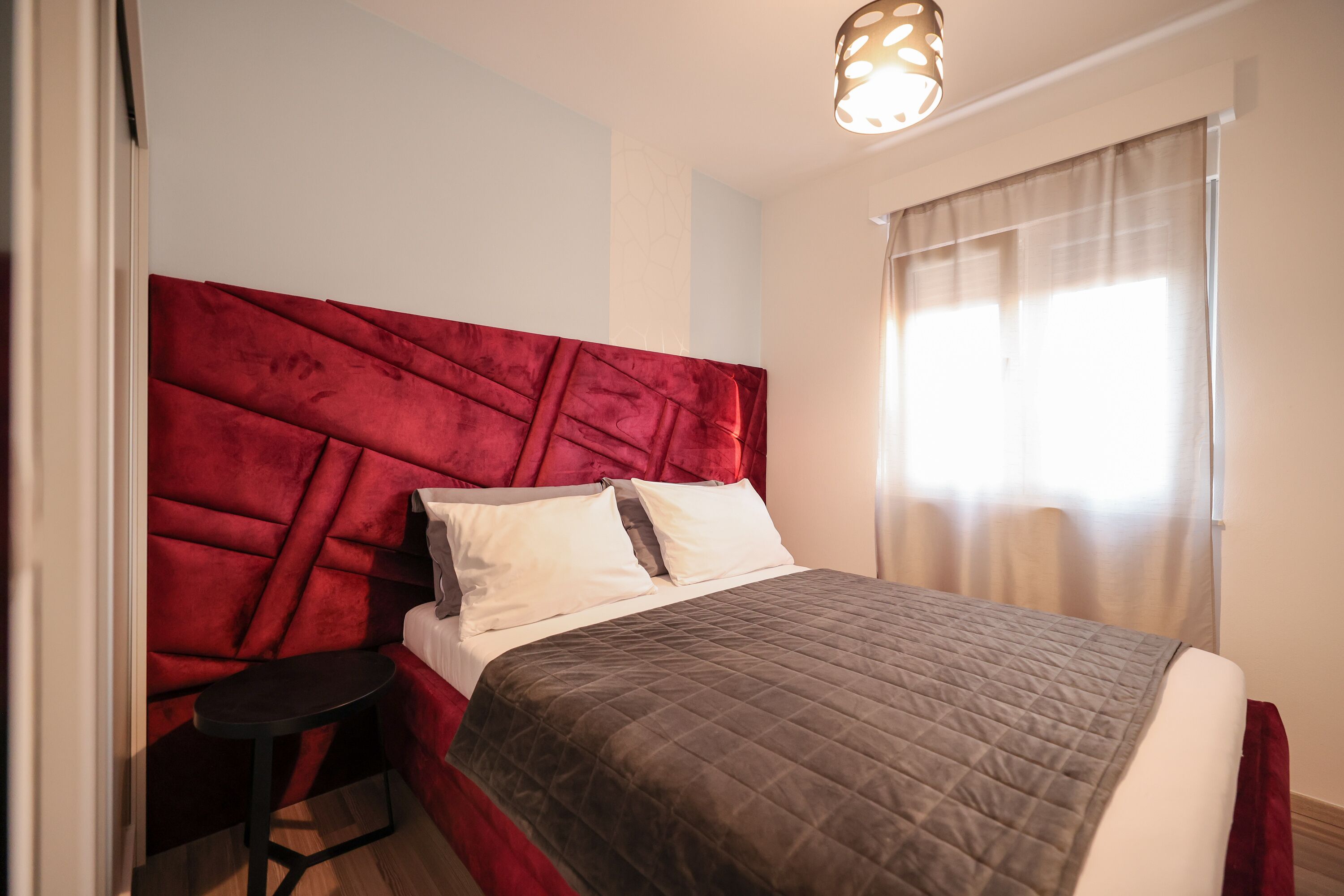 A neatly made bed with a red headboard, a bedside table, and a window with sheer curtains.