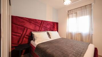 A neatly made bed with a red headboard, a bedside table, and a window with sheer curtains.