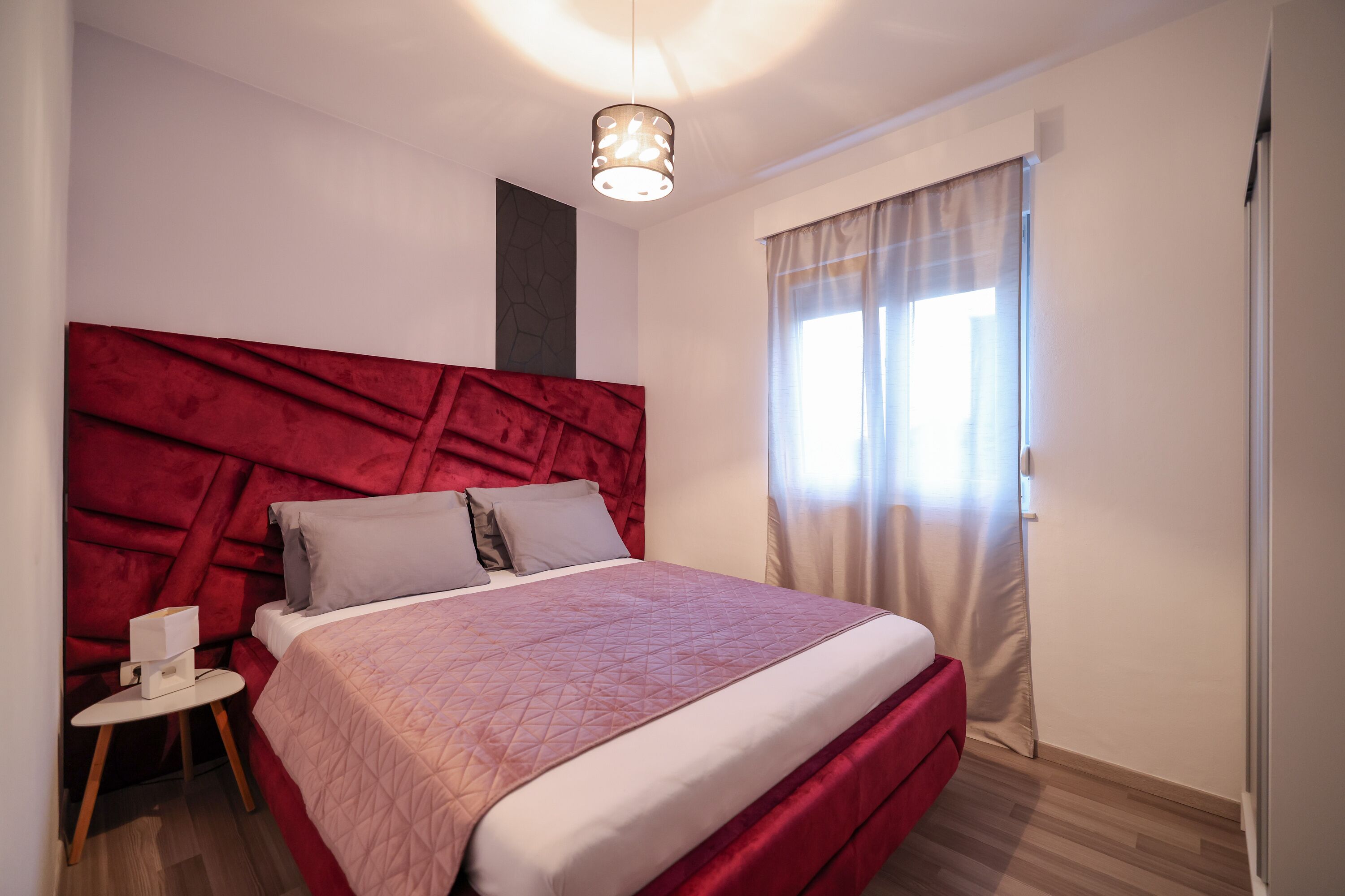 Un dormitori modern amb un capçal de llit vermell, un llit amb llençols roses, una taula de nit amb una làmpada i una finestra amb cortines.