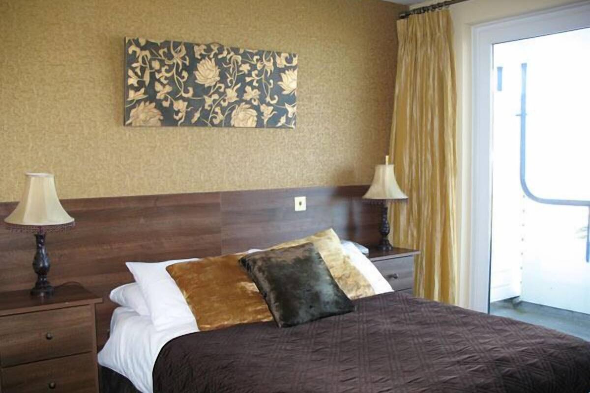 A hotel room with a large bed, two bedside tables with lamps, a floral artwork on the wall, and a view of the outside through a window with curtains.