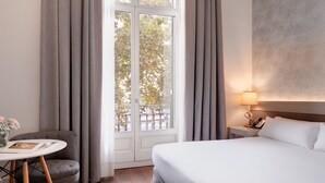 Habitación de hotel con cama, una mesita redonda con un libro y flores, una silla, una mesita de noche con lámpara, y vista a árboles a través de una ventana con cortinas.