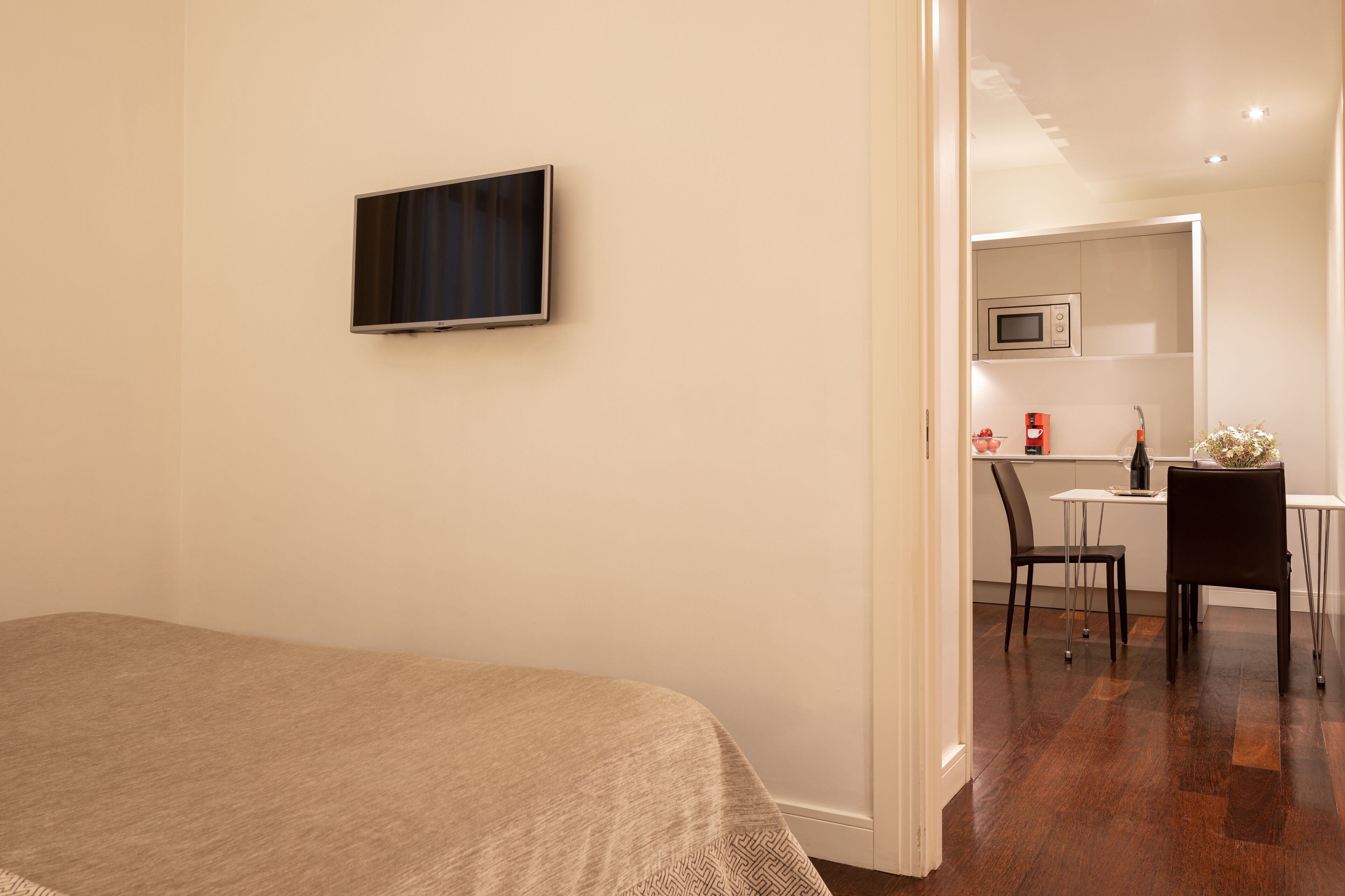 A bedroom with a bed, a TV mounted on the wall, and a kitchenette area with a microwave, a small table, and a chair.