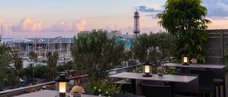 An outdoor dining area with tables and chairs, surrounded by greenery and overlooking a marina with boats and a tower.