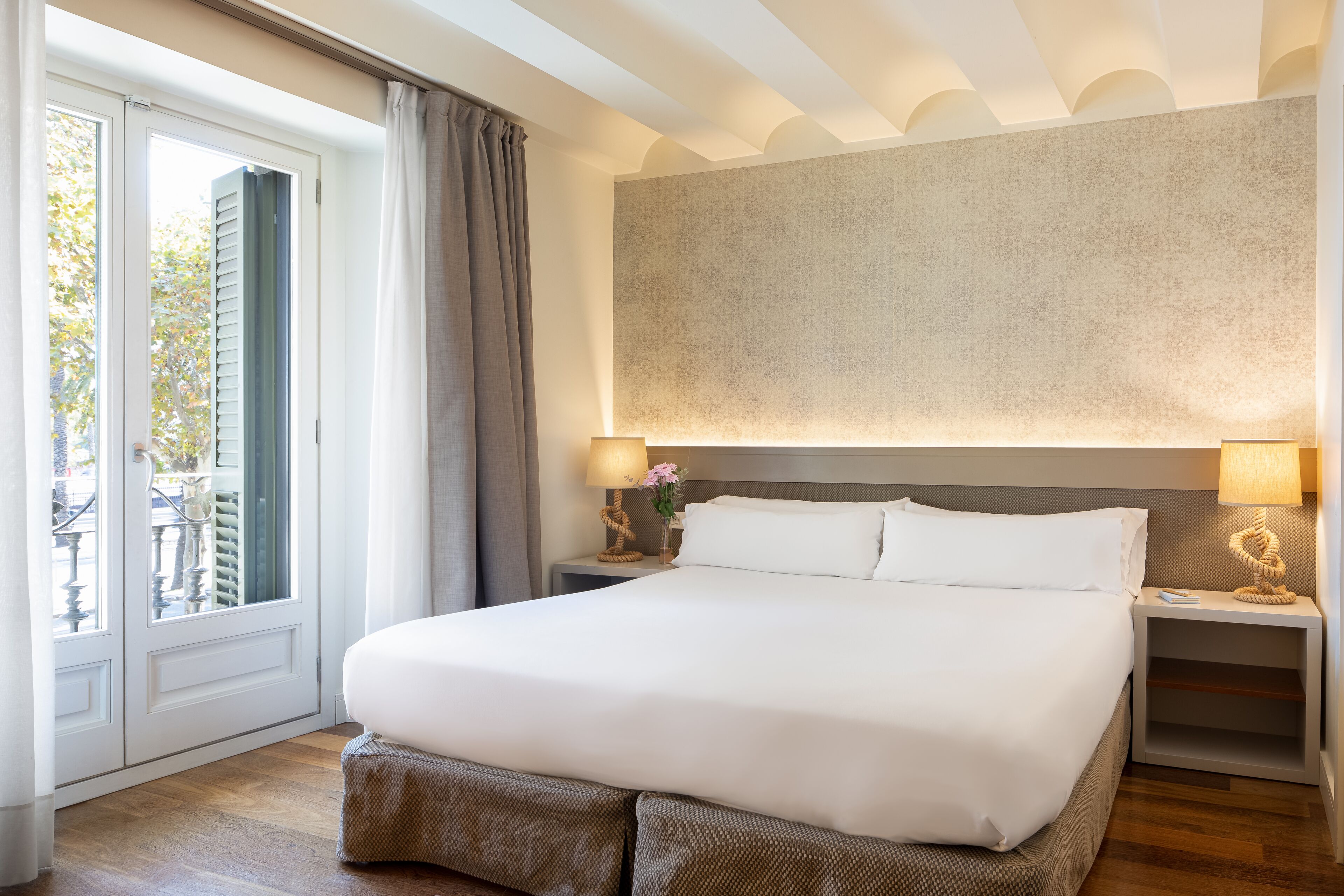 Un lit bien fait dans une chambre d’hôtel, avec une grande fenêtre, une œuvre d’art accrochée au mur et deux lampes de chevet.