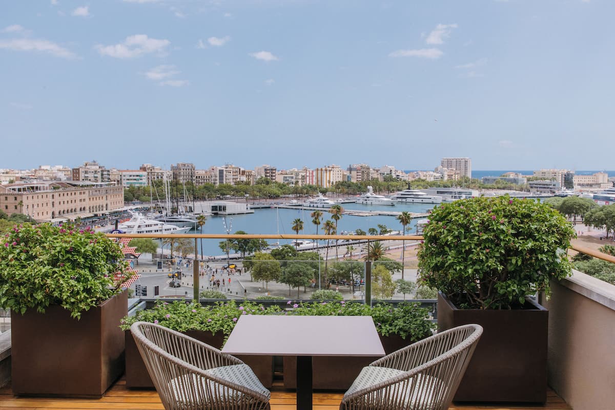 Un balcón con sillas de mimbre y una mesa con vista a un puerto deportivo con barcos y edificios.