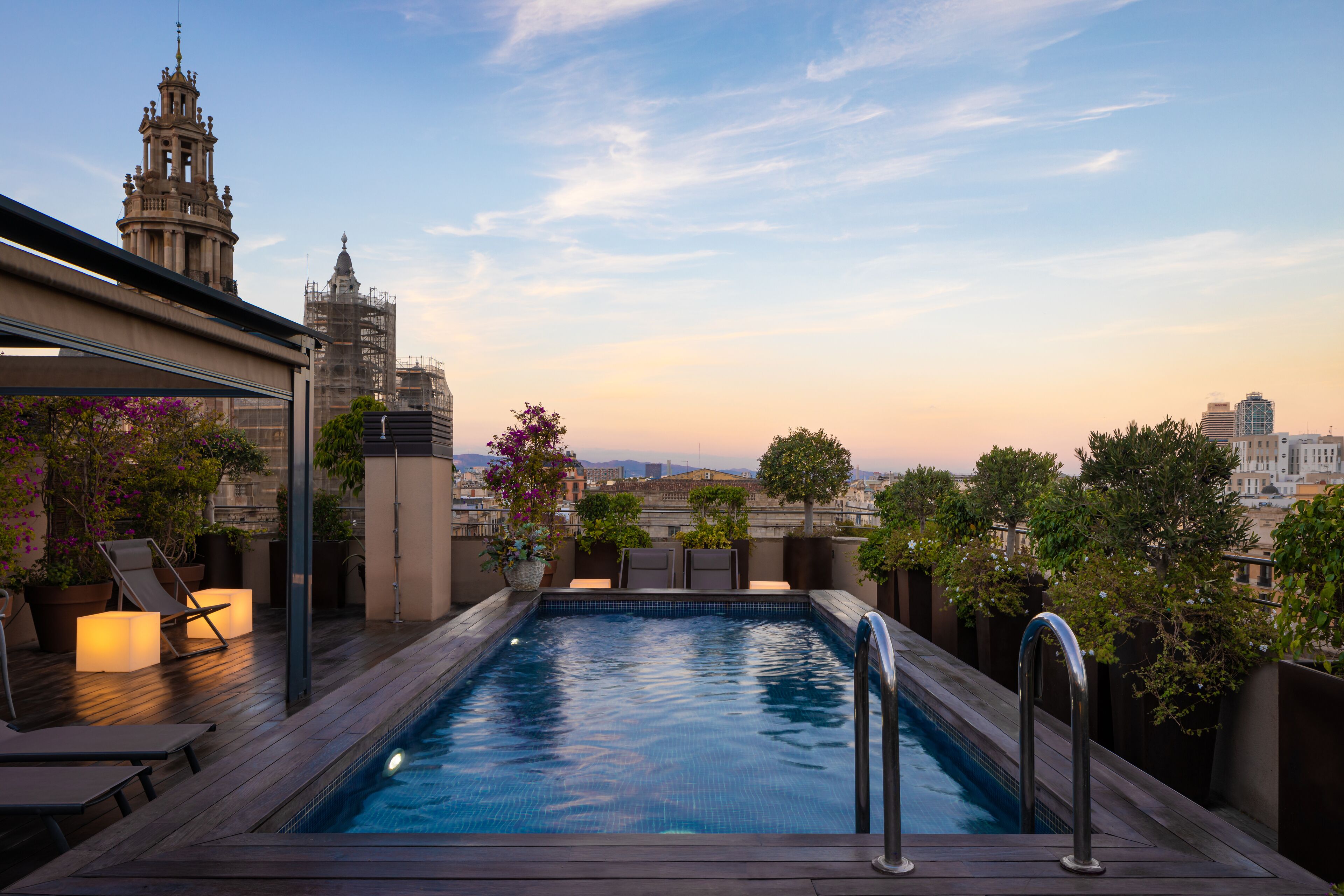 A rooftop pool with a city view, surrounded by lounge chairs and potted plants.