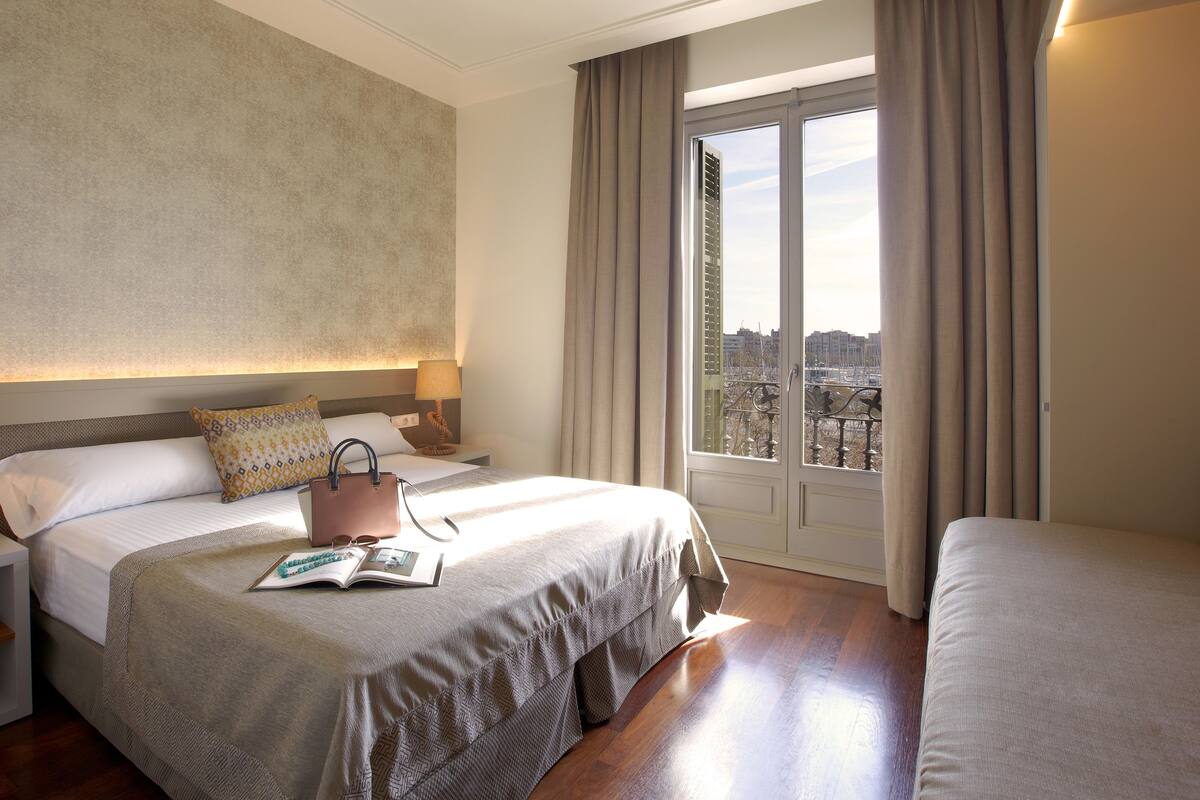 Habitación de hotel con una cama grande, una cama más pequeña, una mesita de noche con lámpara y vistas a la ciudad a través del ventanal.
