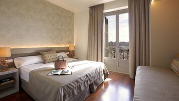 Een hotelkamer met een groot bed, een kleiner bed, een nachtkastje met een lamp, en uitzicht op de stad door het raam.