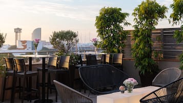 An outdoor seating area with wicker chairs and tables, surrounded by potted plants and overlooking a cityscape.