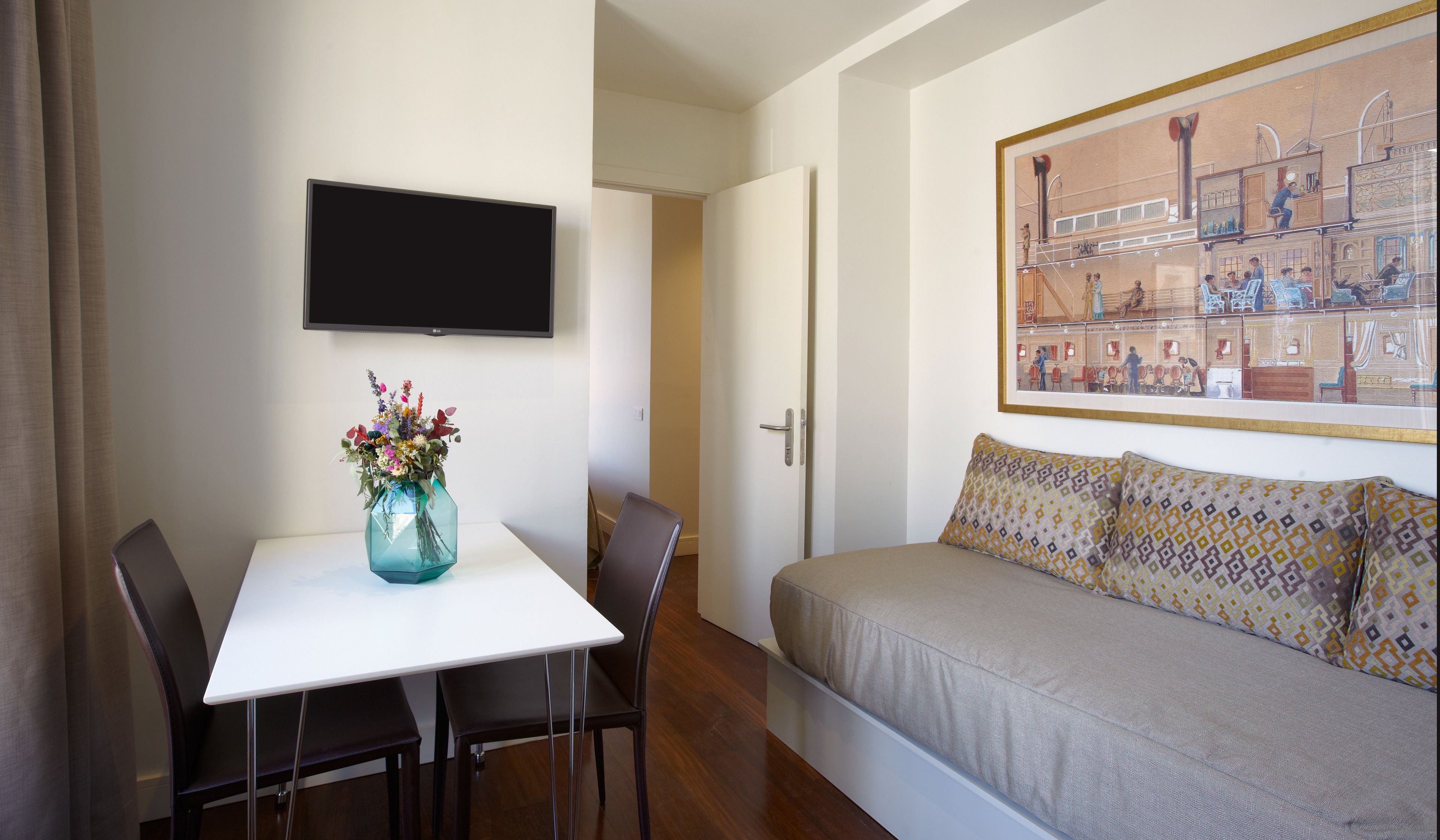 A modern room with a white table, a blue vase with flowers, a sofa with patterned cushions, a flat-screen TV, and a framed picture on the wall.
