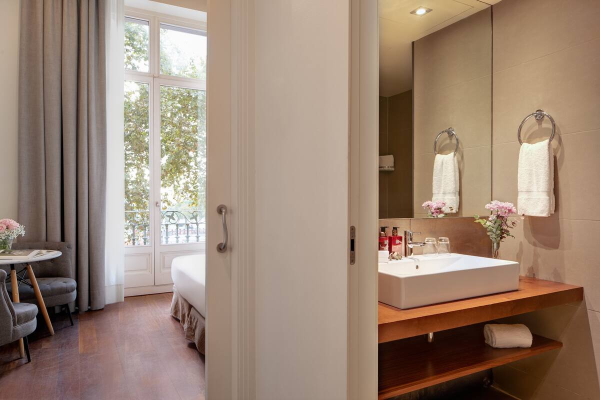 Una habitación de hotel con un ventanal grande, una cama, un tocador de madera con lavabo y un espejo.