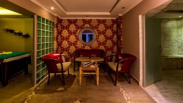 Una habitación con mesa de billar, sillas rojas, una pequeña mesa con revistas y una pared con diseño que tiene un ventanal circular.