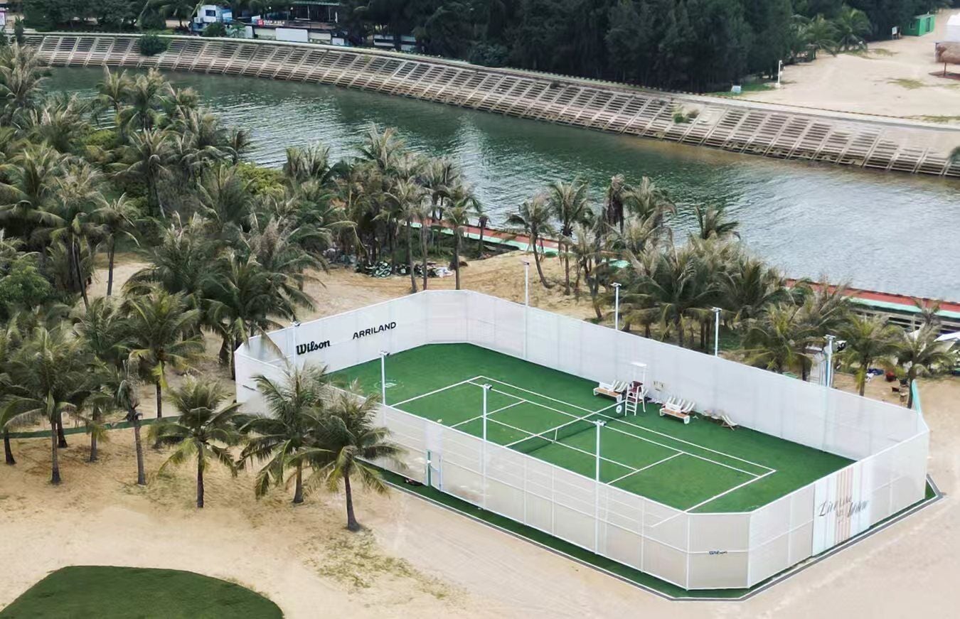 Un court de tennis situé à côté d’une rivière, entouré de palmiers et bordé d’une zone sablonneuse.