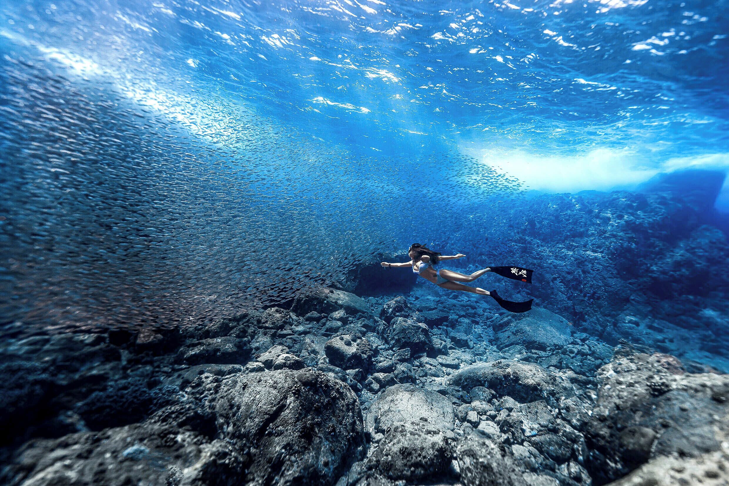 A diver swimming underwater with a rocky seabed and clear water.