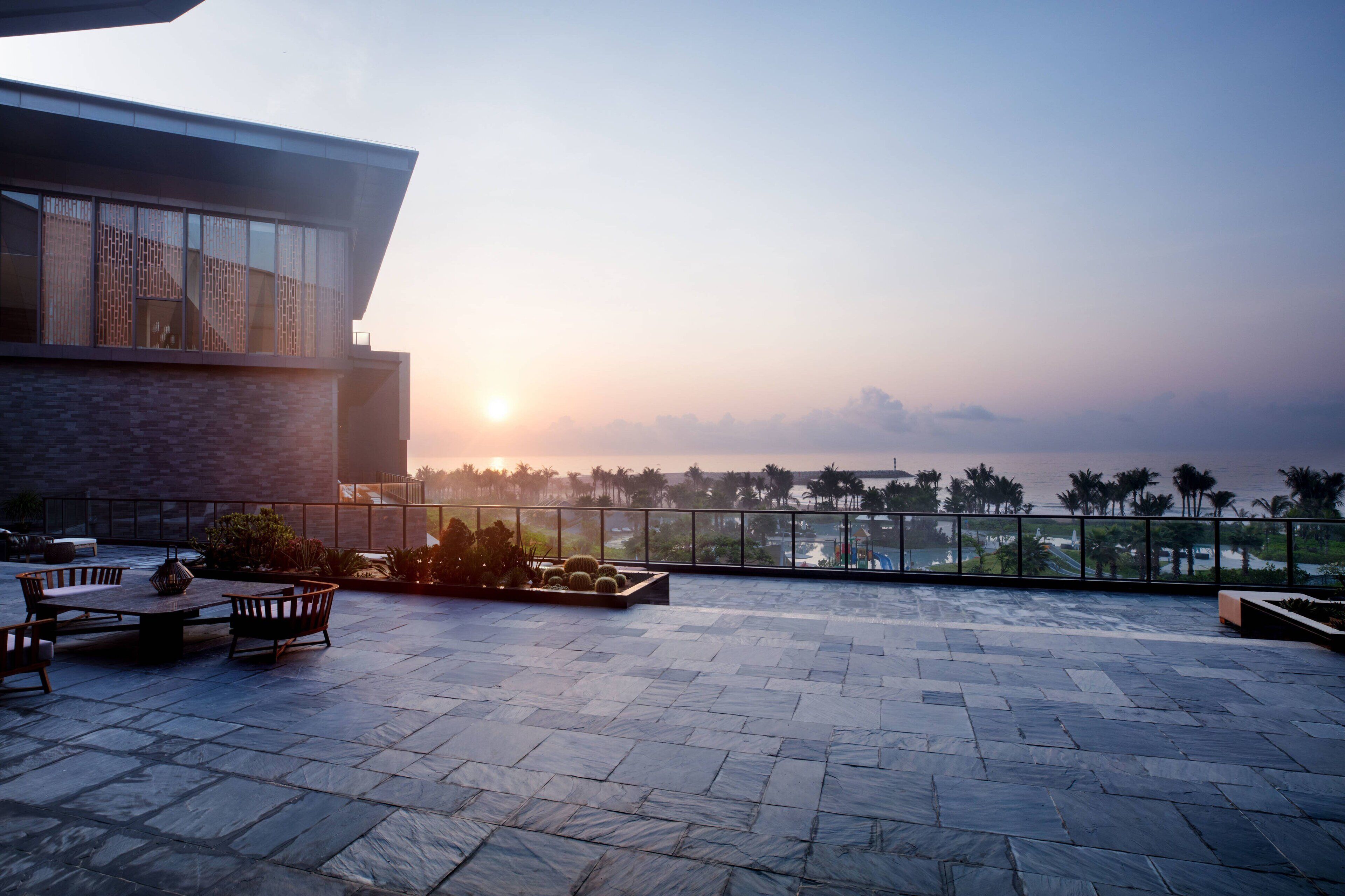 Eine Dachterrasse mit einem modernen Gebäude, Gartenmöbeln und Blick auf den Sonnenuntergang über dem Meer.