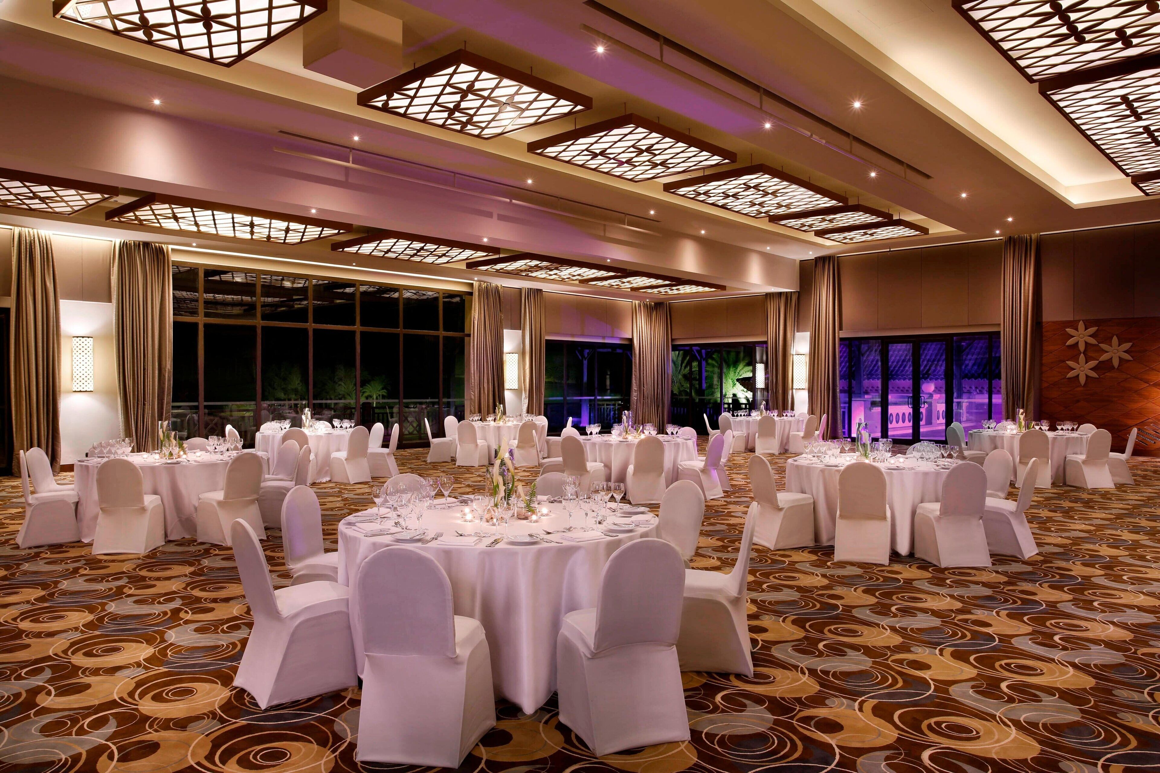 A banquet hall with round tables set for dining, large windows, and a patterned carpet.