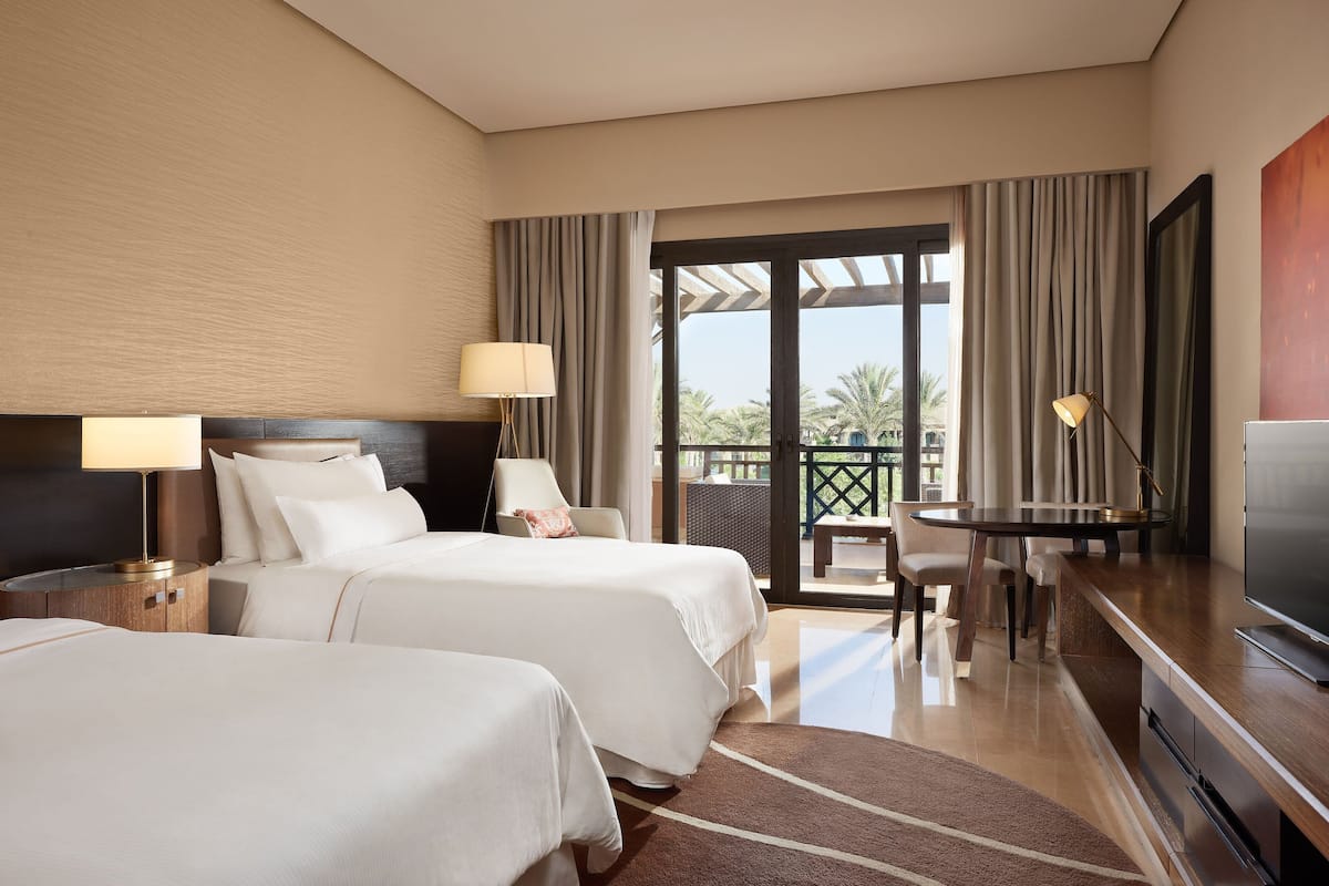 Habitación de hotel con dos camas, un escritorio, televisión y balcón con vistas a palmeras.