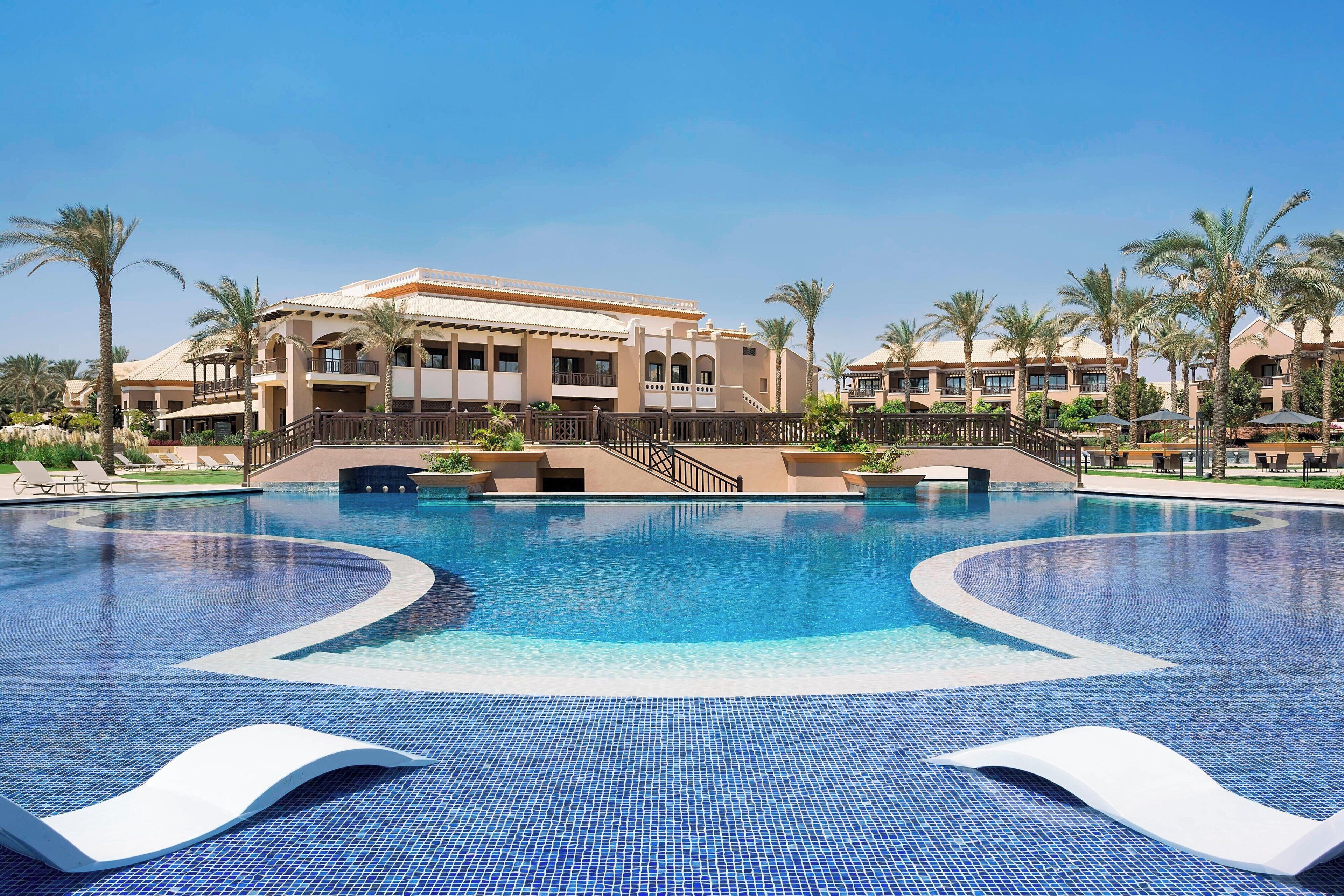 A large, clear swimming pool with a modern design, surrounded by palm trees and a resort building with multiple balconies.