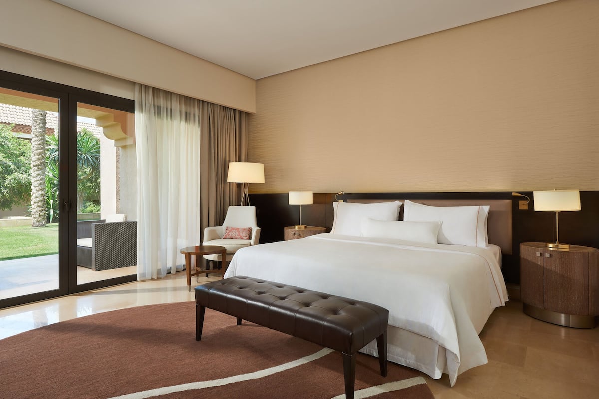 Habitación de hotel con una cama grande, un banco, dos mesitas de noche con lámparas, una silla y vistas al exterior.