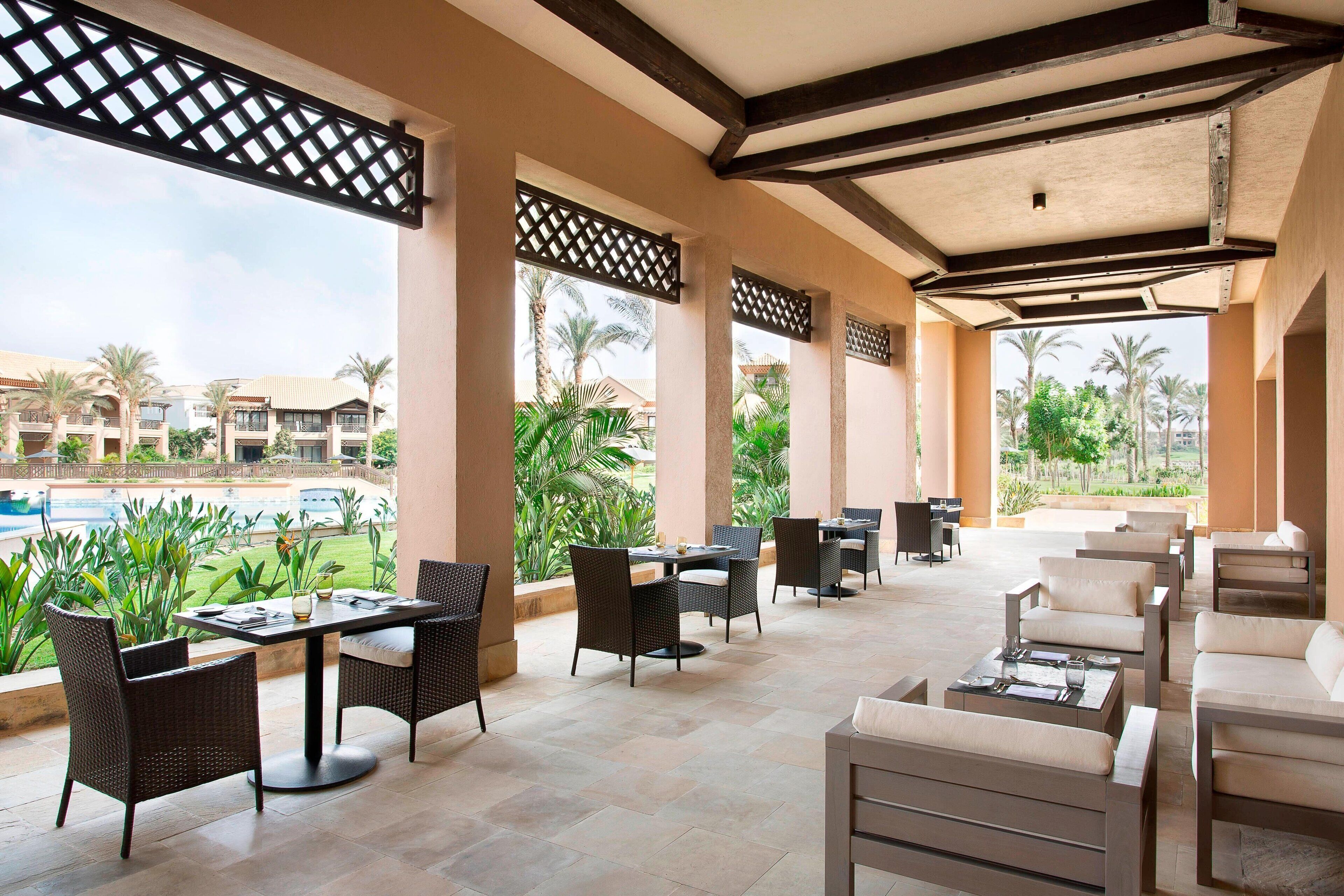 Un espace extérieur aménagé en terrasse, avec des tables et des chaises, de grandes fenêtres et une vue sur une piscine et des palmiers.