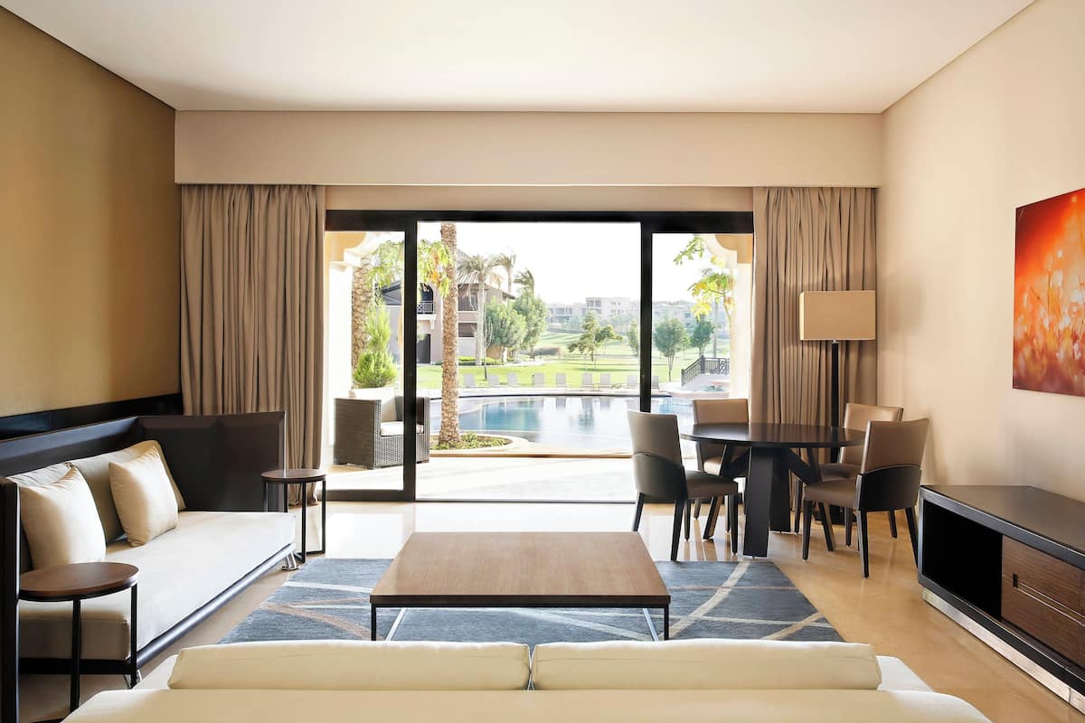 Una habitación de hotel moderna con sofá, mesa de comedor, sillas, televisor y un amplio ventanal con vistas a una piscina y zonas verdes.