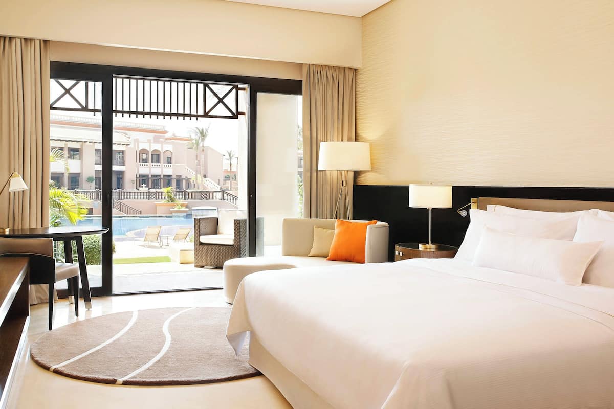 Habitación de hotel con cama, escritorio, silla, lámpara y vista a un área de piscina al aire libre.