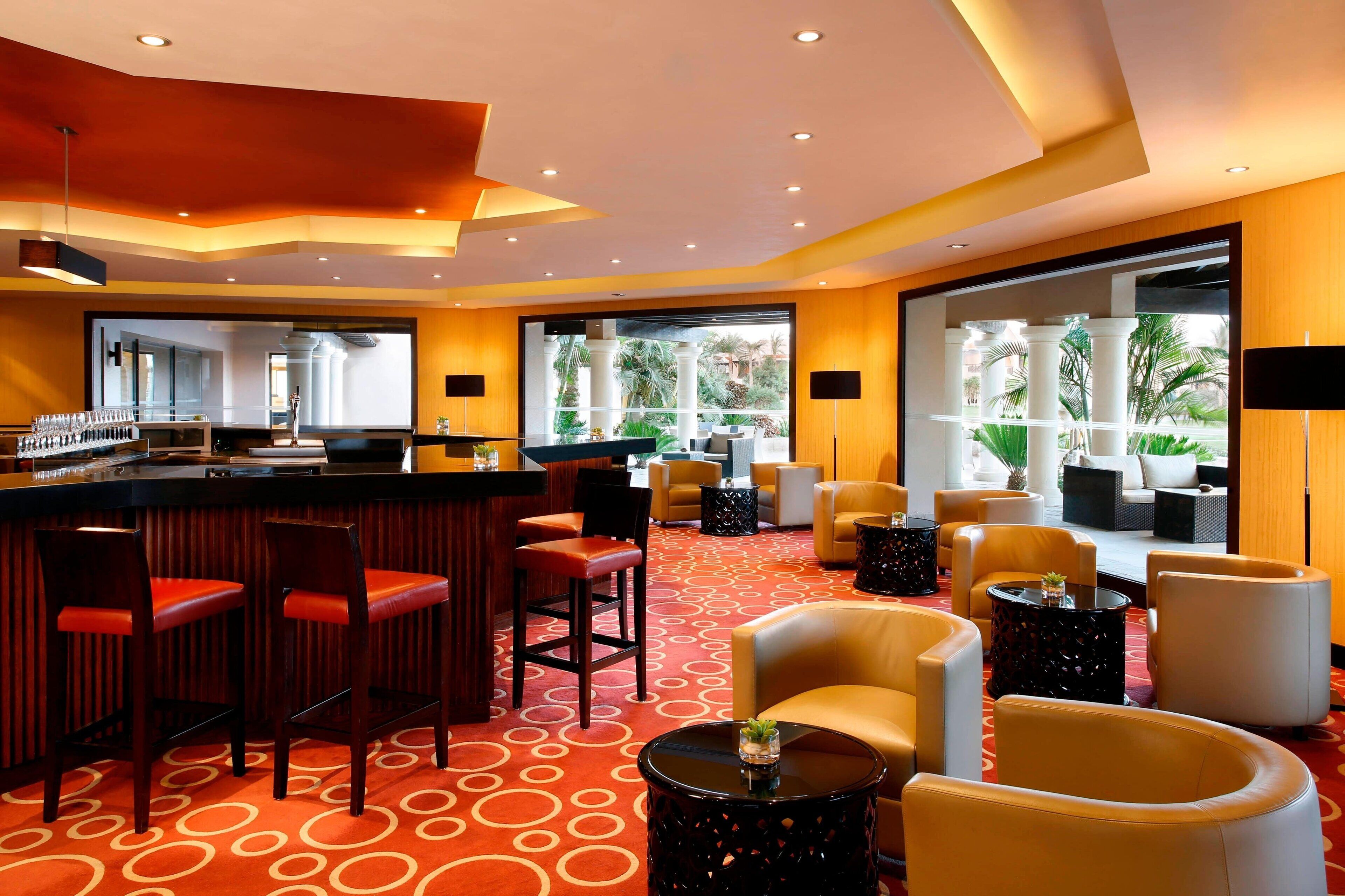 A modern bar area with red bar stools, a counter, and a seating area with yellow chairs and round tables.