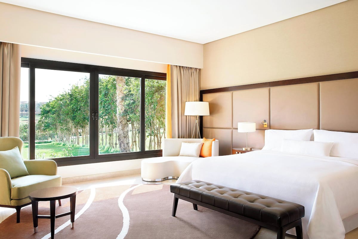 Habitación de hotel con una cama grande, un sofá, una silla y una pequeña mesa. Se ve vegetación a través de la ventana.