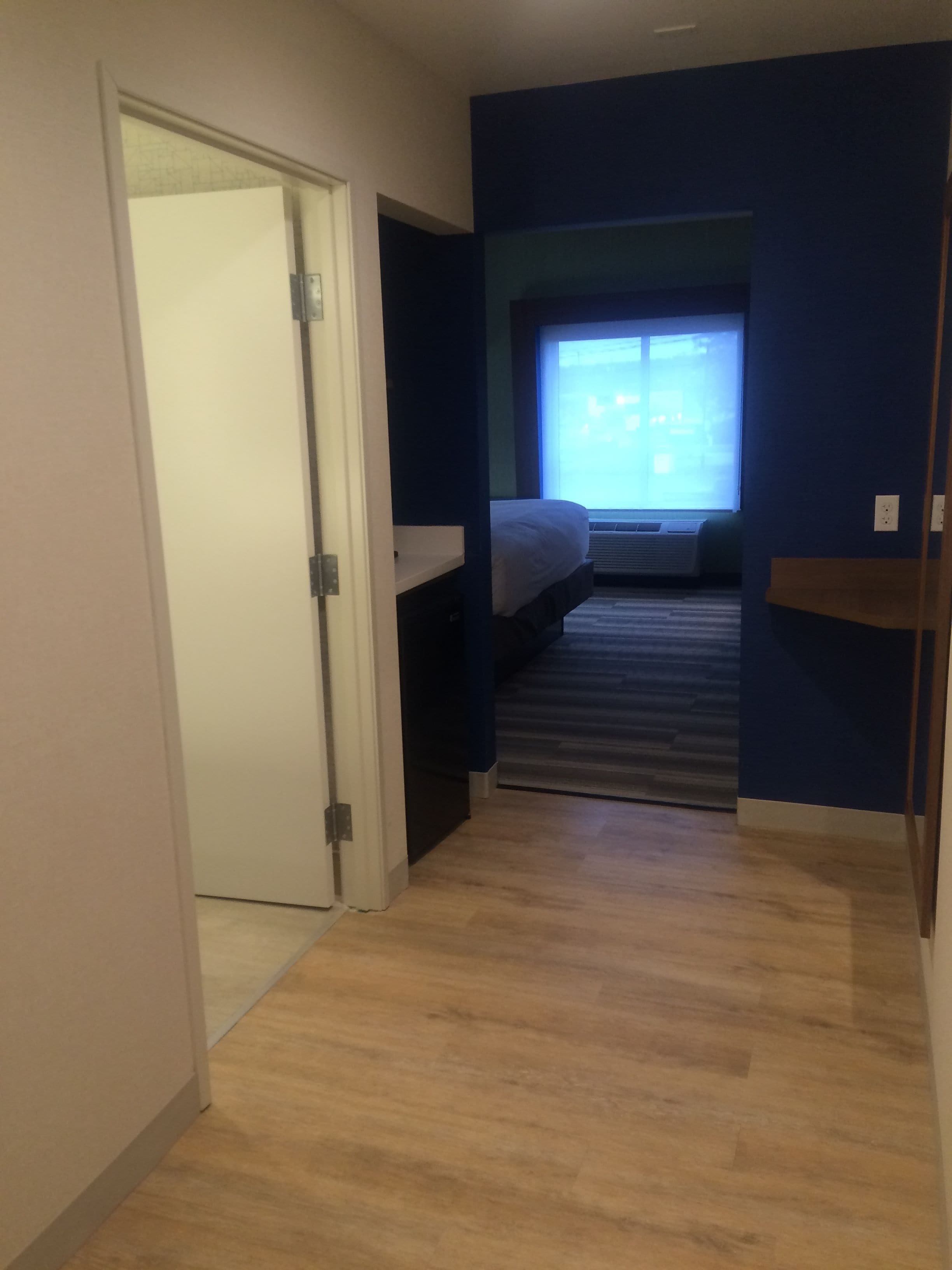 A narrow hallway with a wooden floor leading to a bedroom with a bed and a window.