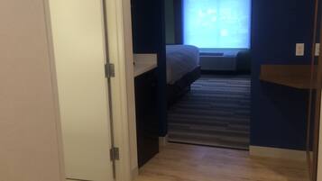 A narrow hallway with a wooden floor leading to a bedroom with a bed and a window.