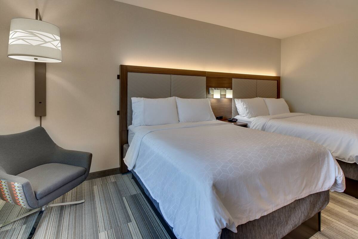 A hotel room with two beds, a gray armchair, and a wall-mounted lamp.
