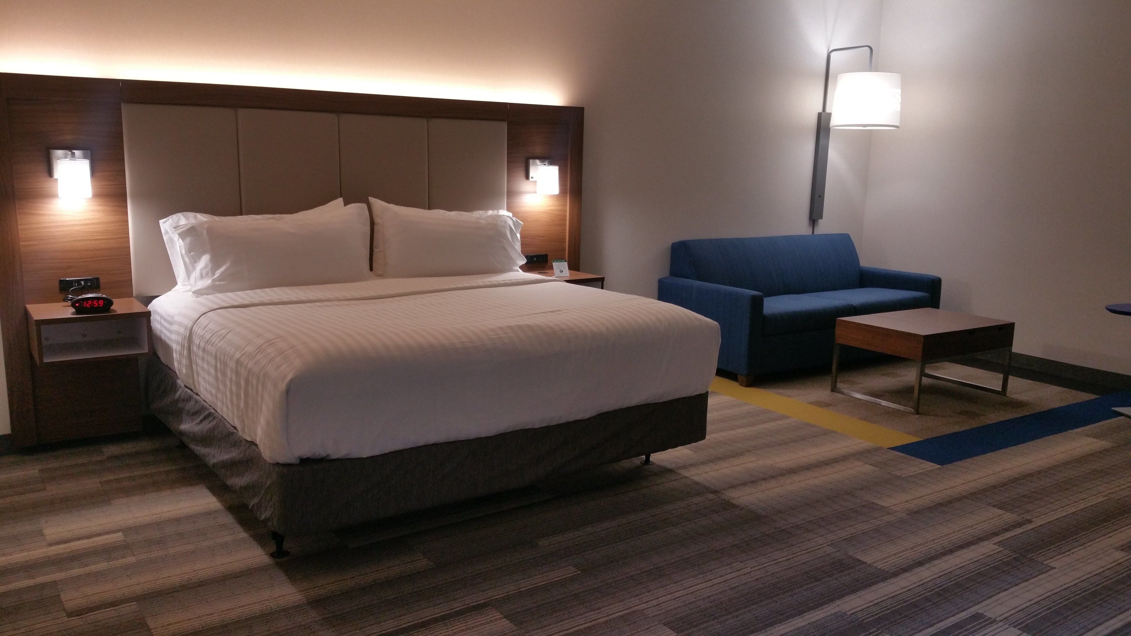 Quarto de hotel com uma cama grande, uma poltrona azul, uma mesinha e uma mesa de cabeceira com um relógio.