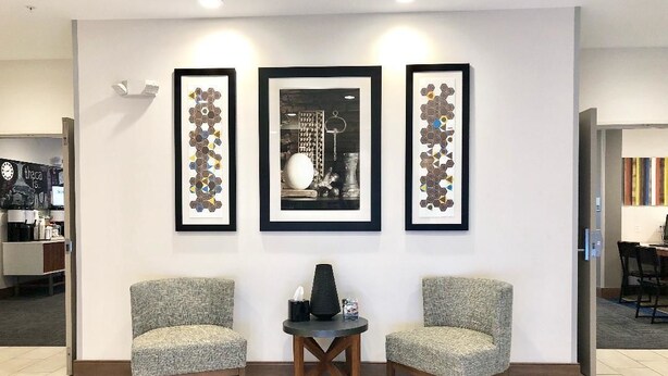 Um lobby com duas poltronas cinzas, uma mesa redonda pequena com um vaso e um livro, e três obras de arte emolduradas na parede.