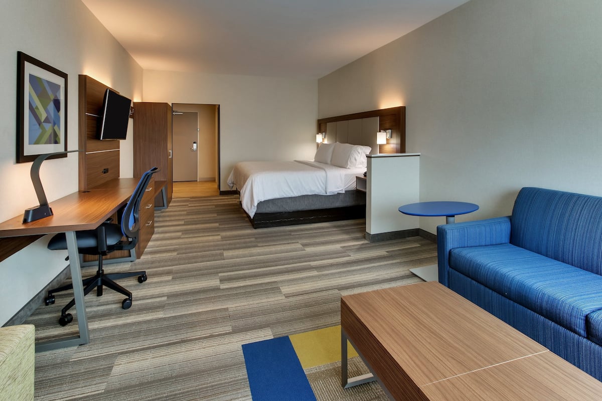 A hotel room with a bed, a desk with a chair, a sofa, a TV mounted on a wooden cabinet, and a small table.